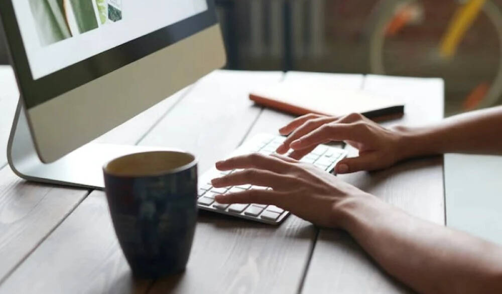 A picture showing hands typing on a keyboard to a computer screen. There's a black  coffee cup and a note book on the table too